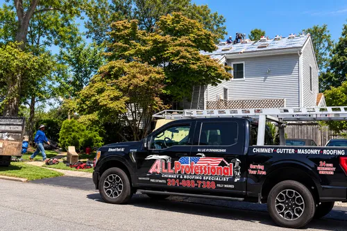 Black ALL Professional pickup with a ladder rack parked on a sunny street while two roofers work on the roof above